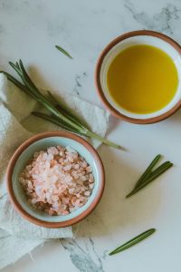 Aesthetic flat lay of Himalayan salt, olive oil, and rosemary on a white marble surface.